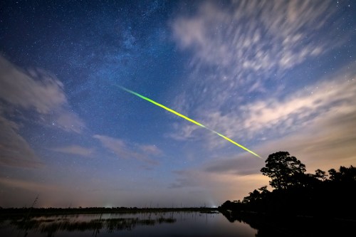 A vibrant green and yellow meteor streaks across a starry night sky over a calm lake and silhouetted trees.