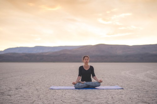 A woman meditates on a yoga mat in a vast, dry landscape at sunset, surrounded by mountains.