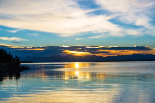 A serene sunset over a calm lake, with colorful reflections on the water and mountains in the background.