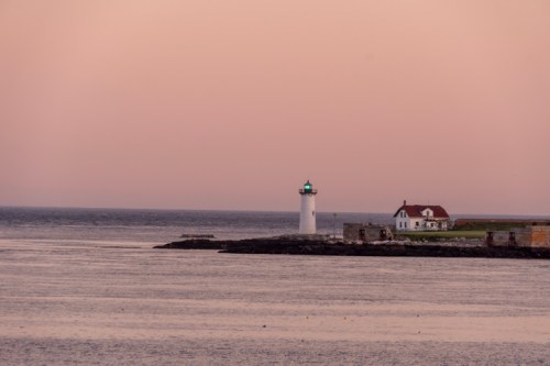A lighthouse stands by the shore at dusk, with a nearby house and calm waters reflecting the soft pink sky.