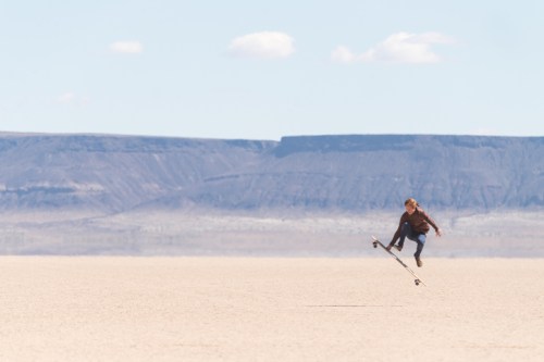 A skateboarder jumps in mid-air over a vast, sandy landscape with distant mountains under a clear blue sky.