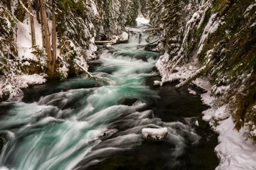 A serene winter scene of a flowing river surrounded by snow-covered trees and rocky banks.