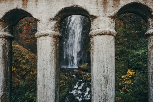 A waterfall cascades through stone arches, surrounded by lush greenery and autumn foliage.