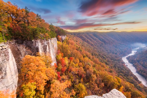 A scenic view of a river winding through vibrant autumn foliage and cliffs under a colorful sky at sunset.