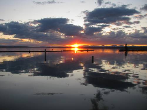 A serene sunset over a calm lake, with vibrant colors reflecting on the water and silhouetted posts in the foreground.