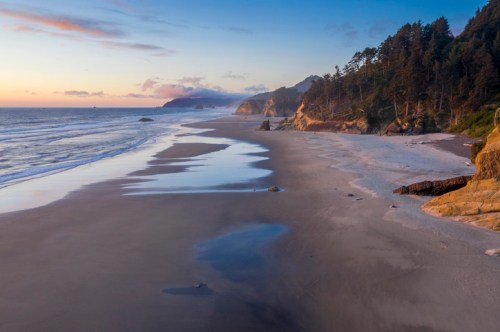 A serene beach at sunset, with gentle waves, sandy shore, and lush trees lining the coastline.
