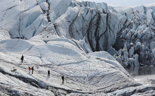 A group of hikers explores a vast, icy glacier landscape with towering ice formations and a calm water body nearby.