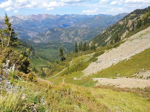 A scenic mountain landscape with rolling hills, green valleys, and distant peaks under a blue sky with fluffy clouds.