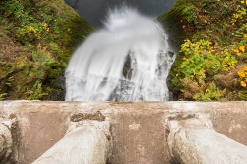 Aerial view of a waterfall cascading over a rocky edge, surrounded by lush greenery and autumn foliage.