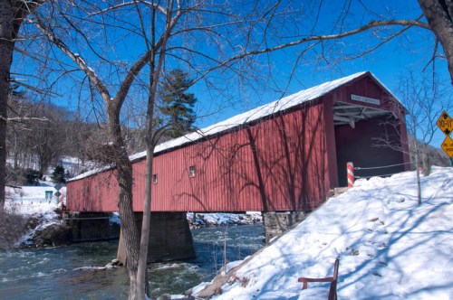 A red covered bridge spans a snowy landscape, with a river flowing beneath and trees in the background.