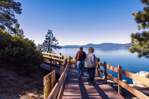 Two people walk along a wooden path by a calm lake, surrounded by trees and mountains in the distance.