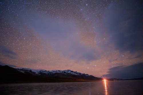A starry night sky over a calm lake, with mountains in the background and a distant light reflecting on the water.