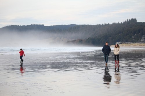 Three people walk along a misty beach, with waves gently lapping at the shore and a forested hillside in the background.