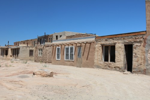 Abandoned adobe buildings under a clear blue sky, with sandy ground and remnants of a once-inhabited area.