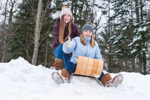 Two girls sit on a sled in the snow, smiling and giving a thumbs up, surrounded by trees in a winter landscape.