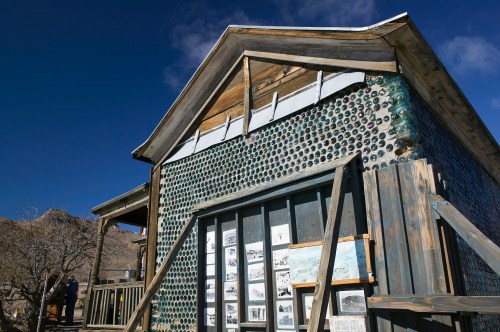 A unique building made of glass bottles, featuring wooden accents and historical photos displayed outside.