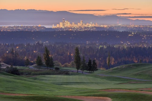 A scenic view of a golf course at dusk, with a city skyline and mountains in the background under a colorful sky.