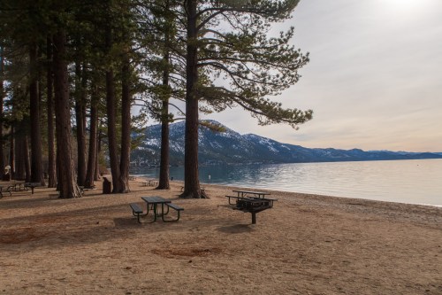 A serene lakeside scene with sandy beach, tall trees, and mountains in the background under a cloudy sky.