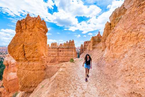 A woman walks along a trail in a canyon with orange rock formations and a blue sky filled with clouds.