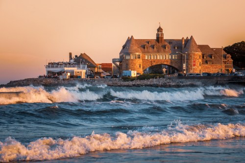 A stone building by the shore at sunset, with waves crashing in the foreground and a boat docked nearby.