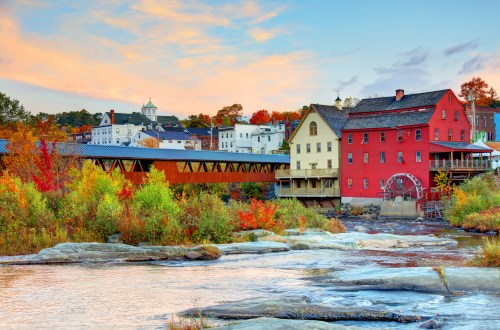 A scenic view of a river with colorful autumn foliage, featuring historic buildings and a covered bridge.
