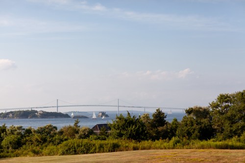 A scenic view of a bridge over water, with sailboats and greenery in the foreground under a clear blue sky.