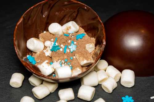 A chocolate bowl filled with marshmallows, sprinkles, and cocoa powder, surrounded by scattered marshmallows.