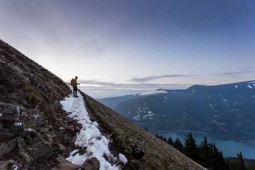 A hiker navigates a snowy trail on a steep mountain slope, overlooking a scenic valley and lake at dusk.