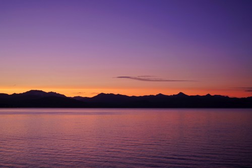 A serene sunset over a calm lake, with silhouetted mountains against a gradient sky of purple and orange hues.