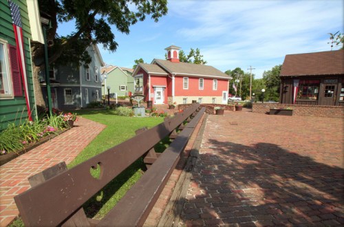 A quaint outdoor area with colorful houses, a red building, and a brick path, surrounded by greenery and flowers.
