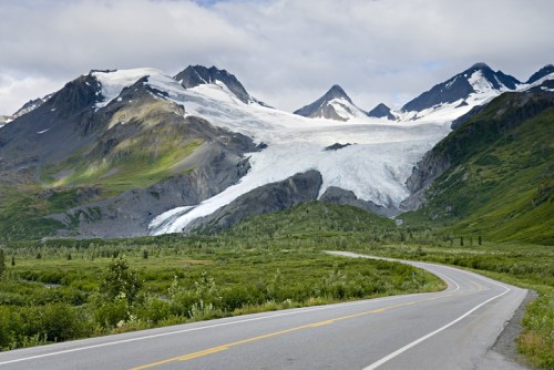 A winding road leads through lush greenery, with majestic mountains and a glacier in the background under a cloudy sky.
