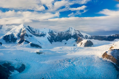 A stunning view of snow-covered mountains and a vast glacier under a blue sky with wispy clouds.