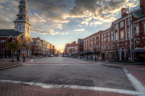 A quiet street scene at sunset, featuring historic buildings and a church steeple under a cloudy sky.