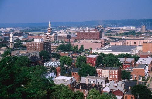 Aerial view of a cityscape featuring buildings, greenery, and a distant industrial area under a clear blue sky.
