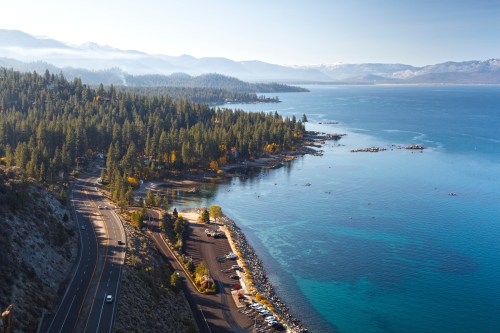 A scenic view of a lake surrounded by mountains and forests, with a winding road and parked cars along the shore.