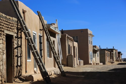 A sunlit street lined with adobe buildings and wooden ladders against a clear blue sky.