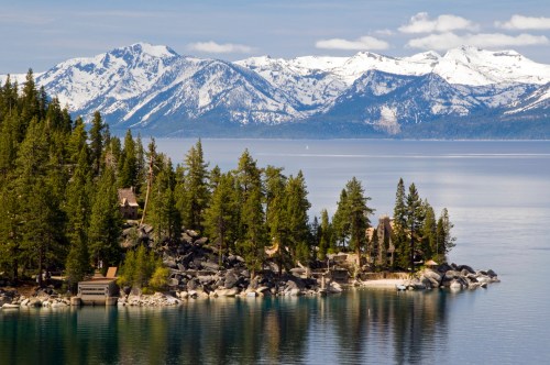 Scenic view of a lake surrounded by snow-capped mountains and lush green trees, with a rocky shoreline.