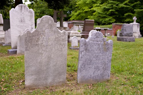 Two weathered gravestones in a grassy cemetery, surrounded by trees and other headstones.