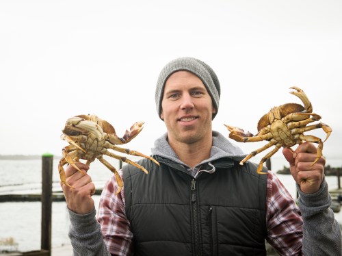A man in a beanie holds a crab in each hand, standing by the water with a cloudy sky in the background.