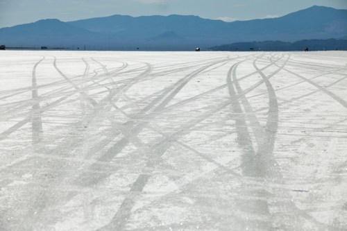 Tire tracks on a vast, white salt flat with distant mountains under a clear blue sky.