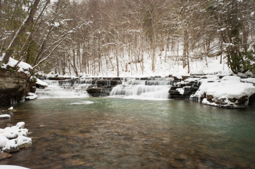 A serene winter scene featuring a snowy landscape with a flowing waterfall and calm, clear water.