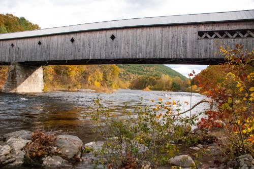 A wooden covered bridge spans a river, surrounded by autumn foliage and rocky riverbanks.