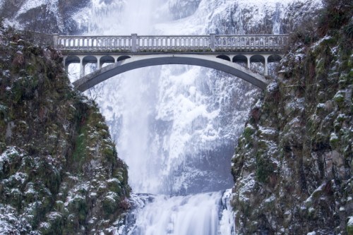 A snowy waterfall cascades beneath a stone bridge, surrounded by lush green cliffs.