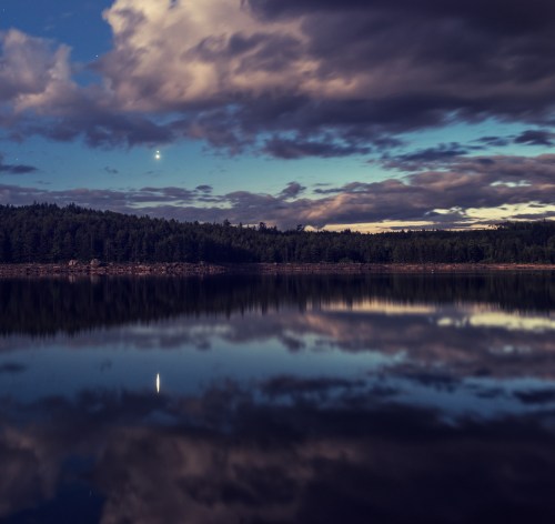 A serene lake reflects a cloudy sky at dusk, with a bright planet visible above the horizon.