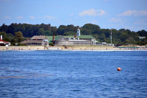 A serene beach scene with a distant view of a building, trees, and a calm blue ocean under a clear sky.