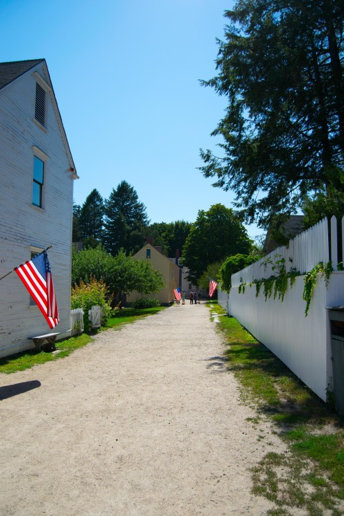 A sunny dirt path lined with white fences and houses, featuring American flags hanging from the buildings.