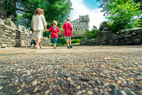 A woman and two children walk along a stone path towards a castle-like structure surrounded by greenery.