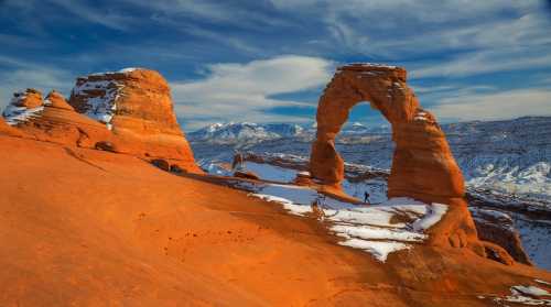 A stunning view of Delicate Arch in Arches National Park, surrounded by snow-capped mountains and a blue sky.