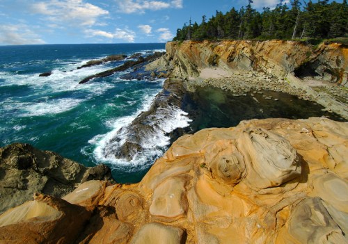 A rocky coastline with waves crashing against cliffs, surrounded by lush green trees under a partly cloudy sky.
