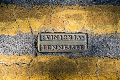 A metal marker on a road, reading "Virginia" and "Tennessee," set against a yellow and gray asphalt background.